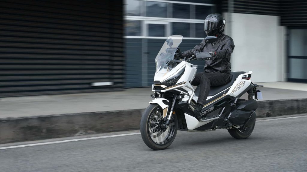 Rider in black protective gear and helmet on a white sport-touring motorcycle cruising down a city street with a tall windshield in clear weather.