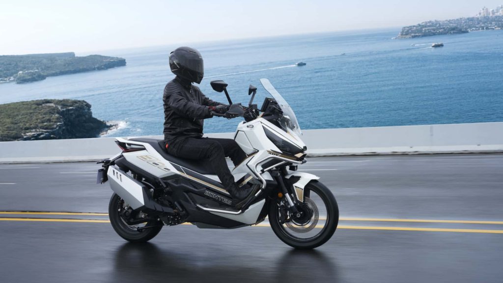 Motorcyclist wearing a full-face helmet and black gear rides a white sport-touring motorcycle along a coastal highway with blue sea in the background